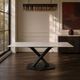 Mesa Cross Statuario con tapa blanca veteada y base negra de patas cruzadas sobre piso de madera.