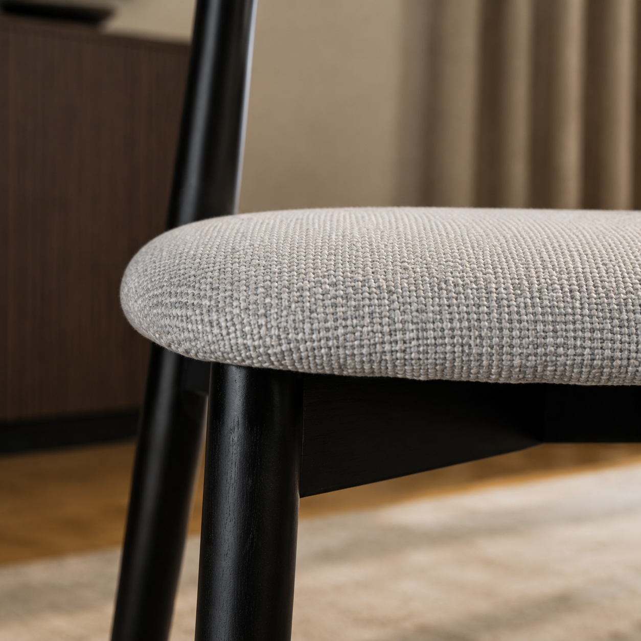 Detalle del borde del asiento tapizado en chenille gris y la terminación de la pata de madera negra de la silla Zuno Deluxe.
