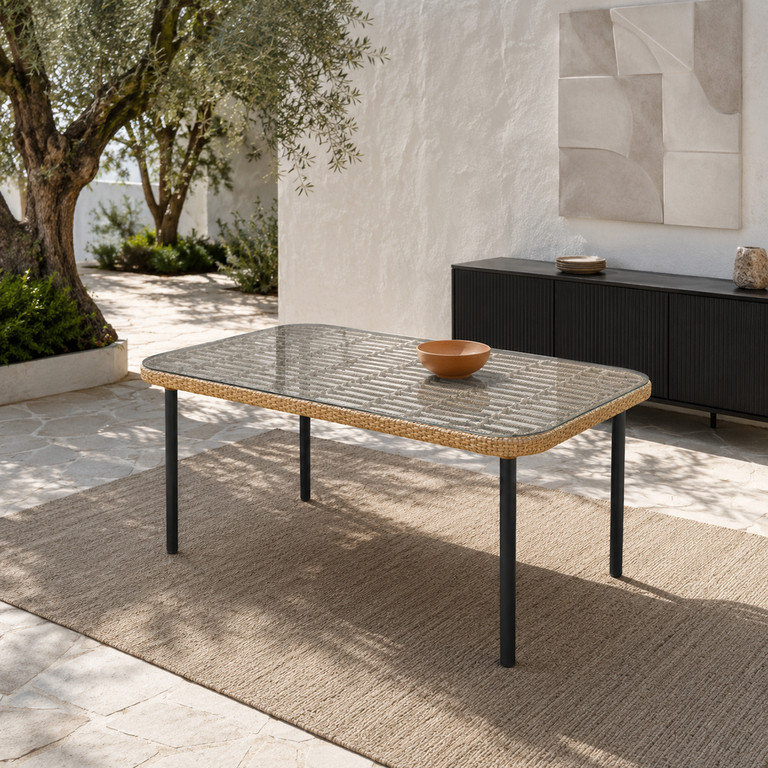 Mesa Exterior Bruce en ratán sintético color natural con tapa de vidrio y patas negras en un patio.